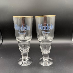May include: Two clear glass Modelo beer glasses with gold rims. The glasses have a unique shape with a flared top and a stem. The word "Modelo" is printed in blue, along with the word "Especial".