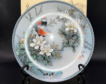 May include: A decorative porcelain plate featuring a detailed painting of two birds perched on a flowering branch. The plate has a light blue background with a detailed border and is displayed on a black stand.
