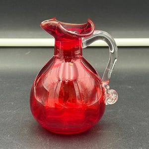 Pode incluir: Uma pequena jarra de vidro vermelho rubi translúcido com um corpo arredondado e facetado e uma alça de vidro transparente. A jarra tem uma borda ondulada e frisada e um bico distinto, exibindo uma cor vermelha vibrante em todo o recipiente.