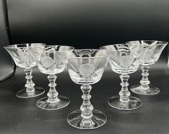 May include: Set of six clear glass coupe glasses. Each glass features a floral design etched into the bowl. The glasses have a short stem and a wide, shallow bowl. The glasses are arranged on a dark surface.