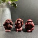 May include: Three red monkey figurines, each with a different gesture: one covering its eyes, one covering its ears, and one covering its mouth. The figurines are sitting on a gray surface.