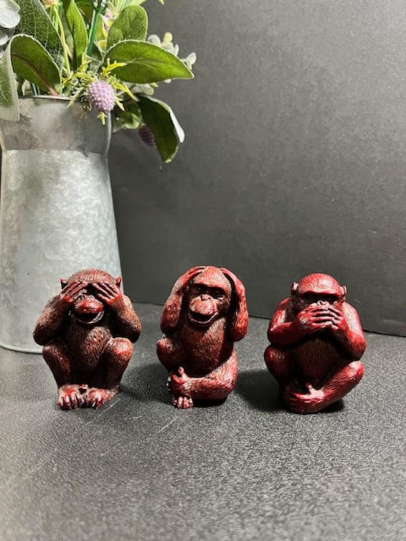 May include: Three red monkey figurines, each with a different gesture: one covering its eyes, one covering its ears, and one covering its mouth. The figurines are sitting on a gray surface.