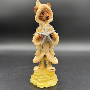 Puede incluir: Una figura de un oso pardo con un abrigo con capucha color crema y una estrella plateada. El oso está sobre una base amarilla. El abrigo está adornado con pequeños adornos azul claro. La cara del oso tiene una nariz negra y ojos oscuros.