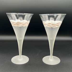 May include: Two champagne flutes with a frosted finish. Each glass has a clear bowl with a decorative band of white and tan woven design near the rim. The flutes have long, slender stems and round bases.