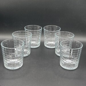 May include: Six clear, short drinking glasses with a textured, grid-like pattern. The glasses are arranged in a group, showcasing their design and clarity. The glassware is suitable for serving beverages.