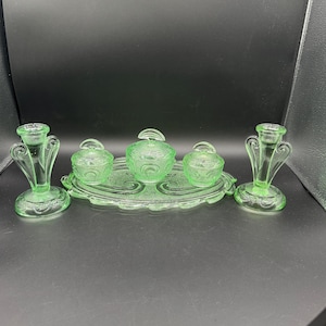 May include: A vintage green glass vanity set, including two candle holders, a serving tray, and three lidded containers. The set features a delicate, etched design and is displayed on a dark surface. The candle holders have a unique, stylized shape.