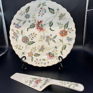 Könnte beinhalten: Ein weißes Keramik-Teller- und Servier-Set mit floralem Design. Der Teller hat einen gewellten Rand und ist mit bunten Blumen und grünen Blättern verziert. Der Servierer hat ein passendes Blumenmuster. Das Set wird auf einem schwarzen Ständer präsentiert.