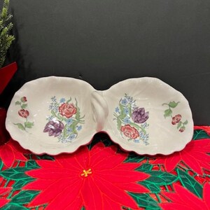 May include: A white ceramic double dish with a floral design. The dish has two separate bowls connected by a handle. The floral design features pink, purple, and blue flowers.