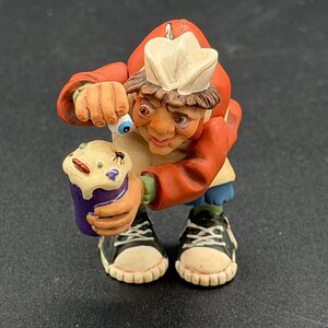 May include: A novelty figurine of a chef holding a cupcake with a bug on top. The chef is wearing a red jacket, white hat, and black trainers. The chef has a surprised expression and is holding an eyeball.
