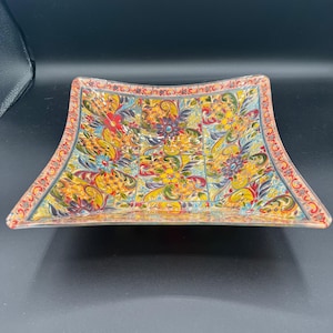 May include: A square decorative dish with a colorful floral pattern. The dish features a vibrant mix of red, yellow, blue, and green hues. The edges are slightly curved, and the overall design is ornate and eye-catching.