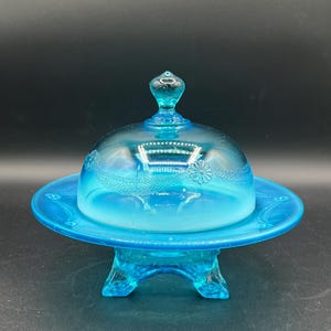 Op de afbeelding: Een vintage, turquoise blauwe glazen botervloot met een gewelfde deksel. Het deksel en de basis hebben reliëf bloemmotieven. De schaal staat op een driepootstandaard en toont een klassiek ontwerp. Het glas heeft een glanzende afwerking.