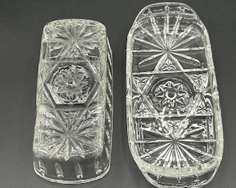 Op de afbeelding: Twee heldere, vintage glazen stukken met ingewikkelde patronen van sterren en geometrische vormen. Eén is een rechthoekige botervloot met een deksel, de andere is een rechthoekige container. Het glas heeft een geslepen kristal uiterlijk.