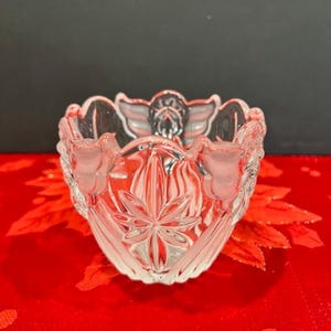 Puede incluir: Un taz&oacute;n de vidrio transparente con un dise&ntilde;o decorativo de &aacute;ngeles y flores. El taz&oacute;n est&aacute; sobre un fondo rojo.