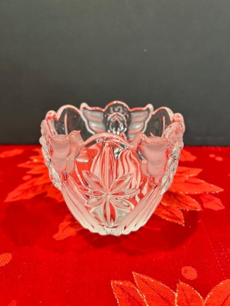 Puede incluir: Un taz&oacute;n de vidrio transparente con un dise&ntilde;o decorativo de &aacute;ngeles y flores. El taz&oacute;n est&aacute; sobre un fondo rojo.