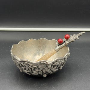 May include: A silver-toned metal bowl with a scalloped rim and a textured leaf pattern on the exterior. A matching metal spoon with a wooden handle and a decorative silver leaf and two red berry accent sits inside the bowl.