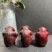 May include: Three red wooden monkey figurines, each with one hand covering their eyes, ears, and mouth. The monkeys are sitting on a dark surface.