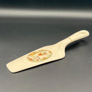 Puede incluir: Servidor de pastel de cerámica blanquecina con un mango decorativo y una hoja ancha y plana. La hoja presenta un diseño ovalado colorido con una escena invernal. El mango tiene un acento dorado.