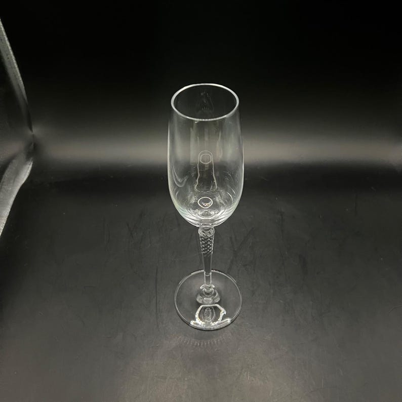 May include: A clear champagne flute with a long stem and a round base. The stem has a textured design. The glass is empty and sits against a black background.