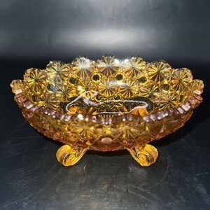 Könnte beinhalten: Bernsteinfarbene Vintage-Glasschale mit Sternmuster und drei kleinen Füßen. Die Schale hat einen gewellten Rand und eine strukturierte Oberfläche, die das Licht reflektiert. Ein dekoratives Stück zum Servieren oder Ausstellen.