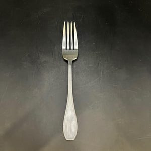 Puede incluir: Un tenedor de acero inoxidable con cuatro púas y un mango decorativo. El mango presenta un patrón vertical texturizado. El tenedor está diseñado para comer y se coloca sobre un fondo oscuro.