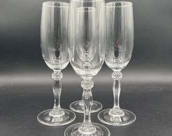 May include: Four clear champagne flutes with a decorative beaded design on the stem. The flutes have a tall, slender shape and sit on a round base. The glasses are made of clear glass and are set against a dark background.