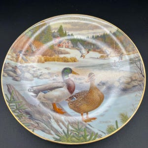 May include: A decorative plate featuring a winter landscape with ducks. The plate has a gold rim and depicts a pair of ducks in the foreground, with other ducks flying in the background. A red house is visible in the distance. The plate is signed "JERNER".