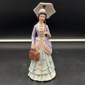 May include: A porcelain figurine of a person wearing a lavender jacket and a light blue skirt with pink rose accents. The figure holds a brown rectangular handbag in one hand and a white parasol over their head, wearing a pink bonnet on their head.