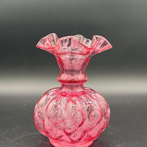 Pode incluir: Um vaso de vidro rosa com uma borda com babados e um corpo bulboso. O vaso tem um padrão texturizado e um pescoço curto. A cor é um rosa vibrante, e o vaso é provavelmente usado para fins decorativos.