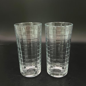 May include: Two clear, tall drinking glasses with a textured, grid-like pattern. The glasses have a cylindrical shape and a thick base. The background is a solid black.