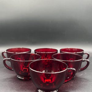 May include: A set of six matching ruby red glass teacups arranged in a cluster. Each cup features a smooth, translucent finish, a rounded bowl shape, a small circular base, and a curved handle extending from the side of the glass vessel.
