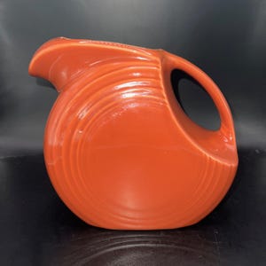 May include: A vibrant orange ceramic pitcher with a rounded body and a curved handle. The pitcher features a spout for pouring and a glossy finish. The design includes vertical ridges that add texture to the surface. This vintage-style pitcher is a kitchenware item.
