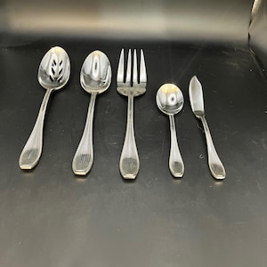 Puede incluir: Un juego de cinco utensilios de servir de color plateado, que incluye una cuchara ranurada, una cuchara de servir, un tenedor, una cuchara pequeña y un cuchillo de mantequilla. Cada pieza tiene un patrón decorativo acanalado en el mango y un acabado pulido.