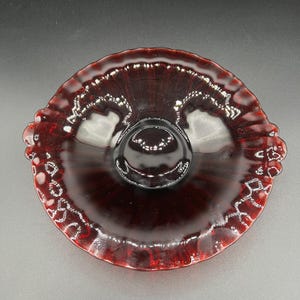 May include: A deep red glass serving dish with a frilled edge and a central ring handle. The dish is circular with a decorative pattern around the rim. The glass reflects light, creating highlights and shadows.