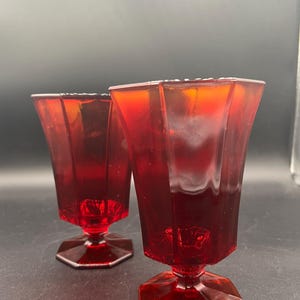 May include: Two matching ruby red glass goblets featuring a faceted, multi-sided design. Each vessel has a wide, flared rim that tapers down to a narrow stem, supported by a solid, hexagonal base. The glass is translucent and deep red in colour.