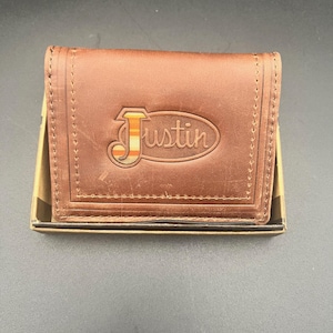 May include: A brown leather wallet with the name "Justin" embossed on the front. The wallet is rectangular and appears to be new, with visible stitching around the edges. The "Justin" logo is in a contrasting color.