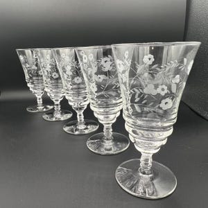Op de afbeelding: Een set van vijf heldere glazen kelken. Elke kelk heeft een geëtst bloemmotief op de kom en een gedetailleerde steel. De kelken zijn in een rij gerangschikt en tonen hun elegante vorm en ingewikkelde details. Ideaal voor het serveren van dranken.