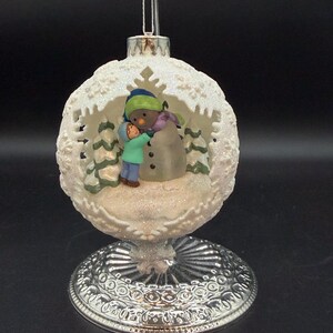 May include: A spherical white ornament with a textured, snowflake-like exterior featuring a cutout window. Inside, a figure in a teal jacket and blue hat hugs a snowman with a green hat and purple scarf. The ornament rests on a silver base.