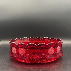 May include: A round, ruby red glass bowl featuring a scalloped rim and a series of embossed circular medallions around the exterior. The medallions display alternating designs of the Liberty Bell and a profile portrait, set against a sunburst pattern.
