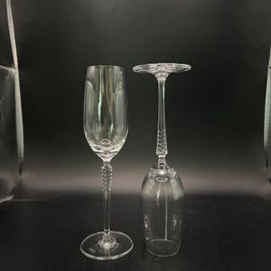 May include: Two clear crystal champagne flutes. One is upright, the other is inverted. Both have a long, twisted stem and a round base. The flutes are set against a dark background.