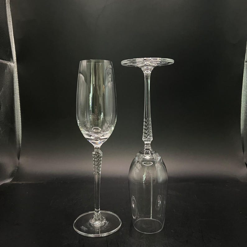 May include: Two clear crystal champagne flutes. One is upright, the other is inverted. Both have a long, twisted stem and a round base. The flutes are set against a dark background.