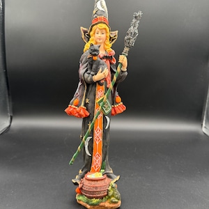 Könnte beinhalten: Figur einer Hexe, die eine schwarze Katze und einen Besen hält. Die Hexe trägt ein schwarzes Kleid mit orangefarbenen Verzierungen, einen hohen Hut mit einem Mond und hat blondes Haar. Ein Kessel und ein Frosch befinden sich am Boden.