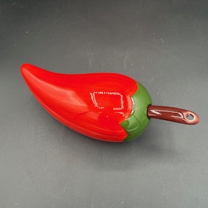 Pode incluir: Um utensílio de cozinha em cerâmica em forma de pimenta, principalmente vermelho com um caule verde e uma alça marrom. A alça tem um buraco na extremidade. A superfície brilhante reflete a luz.