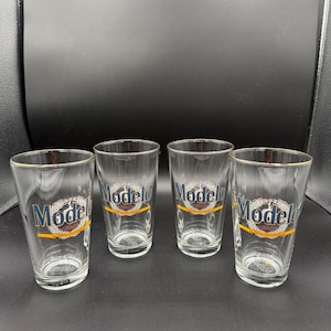 May include: Four clear pint glasses with the Modelo logo in blue, gold, and white. The glasses have a gold rim and are arranged in a row. The logo is printed on the front of each glass.