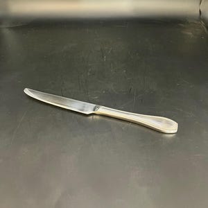 Puede incluir: Un cuchillo de mantequilla plateado con un mango curvo y una hoja dentada. El mango tiene un diseño estriado. El cuchillo está colocado horizontalmente sobre una superficie oscura.