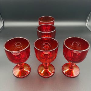 May include: A set of six red glass goblets with a silver rim. The goblets have a rounded bowl shape and sit on a short stem and a circular base. The glass has a glossy finish, reflecting light.