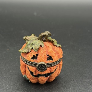 May include: A small, orange pumpkin-shaped trinket box with a smiling jack-o'-lantern face. The box has a dark metal band around the middle and green leaves on top. The box is a decorative item.