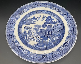 Może przedstawiać: Okrągły, biało-niebieski ceramiczny talerz z klasycznym wzorem Willow. Talerz przedstawia centralną scenę z pagodą, łodzią i ptakami, otoczoną szczegółową obwódką. Talerz ma średnicę około 25 cm.