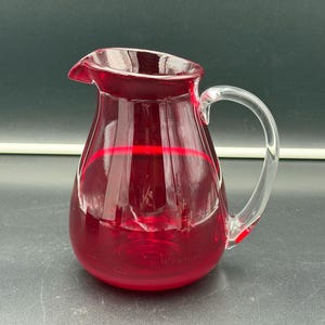 Puede incluir: Una jarra de cristal rojo rubí translúcido con cuerpo redondeado, pico vertedor ancho y un asa de cristal transparente curvada unida al lateral. El recipiente tiene una superficie lisa y pulida que refleja la luz, mostrando su color.