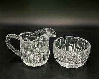 Op de afbeelding: Set van een heldere glazen roomkannetje en suikerpot. Het kannetje heeft een handvat en een tuit, terwijl de suikerpot rond is. Beide stukken hebben een gedetailleerd, reliëfpatroon. De set is ontworpen om te serveren.