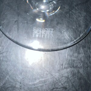 May include: A clear glass with the text "SCHOTT ZWIESEL" etched into the glass. The glass has a curved rim and is partially visible against a reflective, textured surface.